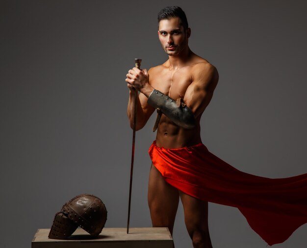 Muscular man ancient Rome soldier holding a sword dressed in a red fluttering dress.
