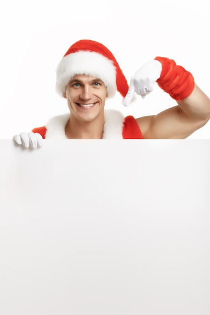 Foto gratuita uomo muscolare travestito da babbo natale che punta a un cartellone bianco mentre sorridendo