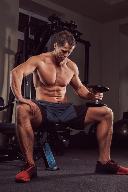 A muscular shirtless athlete doing exercise with dumbbells while sits on a bench in the gym.