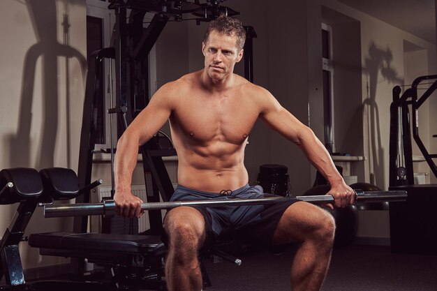 Мускулистый спортсмен без рубашки делает упражнения с гантелями, сидя на скамейке в тренажерном зале.