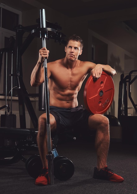 A muscular shirtless athlete holds barbell disc while sits on a bench in the gym.