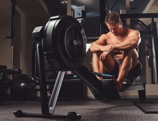 근육질의 셔츠를 입지 않은 운동선수가 체육관에서 로잉 머신에 앉아 있는 동안 힘든 운동 후에 쉬고 있습니다.