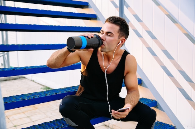 Muscular strong man drinking a water after training, listening to music in earphones