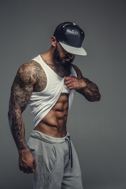 Muscular tattooed man with beard looking down and posing on camera. Isolated on grey background.