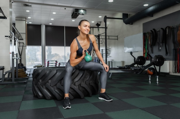 Muscular woman trains biceps with kettlebell in her hands while sitting on large wheel in the gym. fitness concept