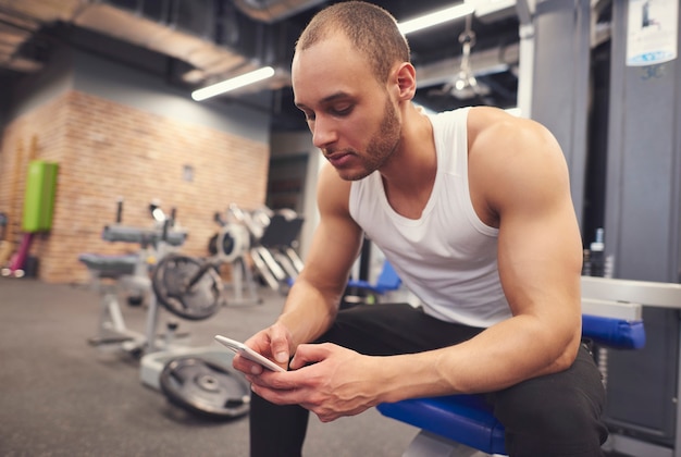 Free photo muscular young man using cell phone at gym