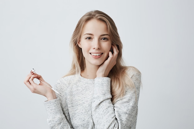 Music, happiness and technology concept. Lovely beautiful female wears her long blonde hair loose with white earphones, listens to music on cell phone, uses music app, poses against gray wall.