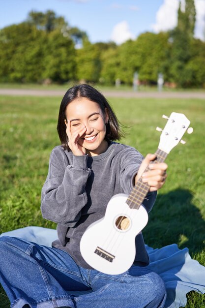 音楽と楽器笑顔のアジアの女の子は彼女の白いウクレレが公園に座っていることを示し、小さなギターを弾きます