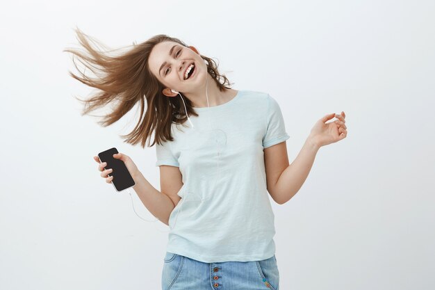 Music is great emotion booster. Portrait of charming joyful and emotive woman jumping happily waving hair and smiling from joy listening music in earphones holding smartphone posing against grey wall