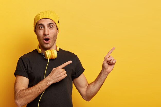 Music is part of technology. Surprised Caucasian man wears headphones, yellow headgear and black t shirt