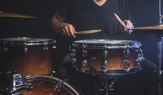 A musician in a black T-shirt plays drums with sticks on stage.