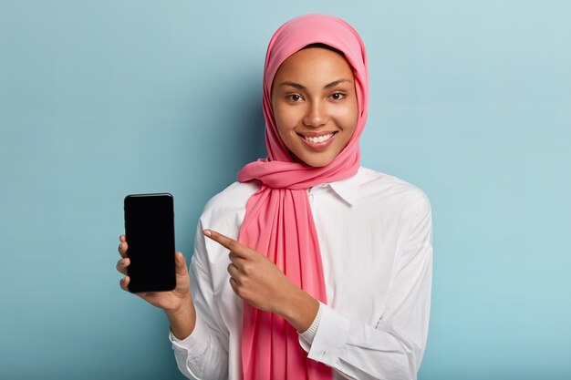 Muslim lady holds smartphone, shows blank screen to insert text or your information, wears pink hijab and white shirt