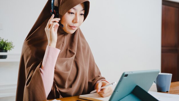 イスラム教徒の女性がデジタルタブレットを使用してヘッドフォンを着用し、自宅のキッチンで仕事をしているときに、会議のビデオ通話で販売レポートについて同僚に話します。