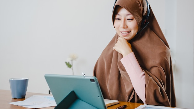 부엌에서 집에서 일하는 동안 회의 화상 통화에서 판매 보고서에 대해 동료에게 디지털 태블릿을 사용하는 이슬람 여성 착용 헤드폰.