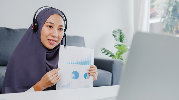 랩톱을 사용하는 이슬람 여성 착용 헤드폰은 거실에서 집에서 일하는 동안 회의 화상 통화에서 판매 보고서에 대해 동료에게 이야기합니다.