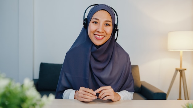 밤에 재택 근무하는 동안 회의 화상 통화에서 판매 보고서에 대해 동료에게 랩톱을 사용하는 이슬람 여성 착용 헤드폰.