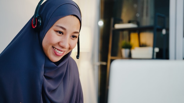 イスラム教徒の女性がヘッドフォンを着用するウェビナーオンラインコースを聞く夜のホームオフィスでの会議ビデオ通話で通信します。