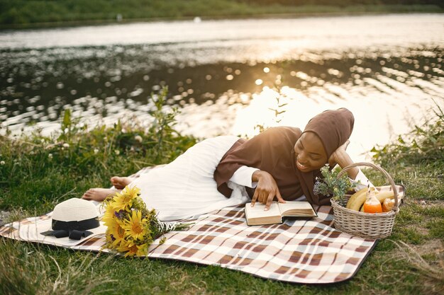 한 이슬람 여성이 강 근처의 격자 무늬 피크닉 담요에 누워 책을 읽고 있다