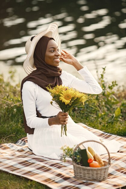 이슬람 여성이 강 근처의 격자 무늬 피크닉 담요에 앉아 있다