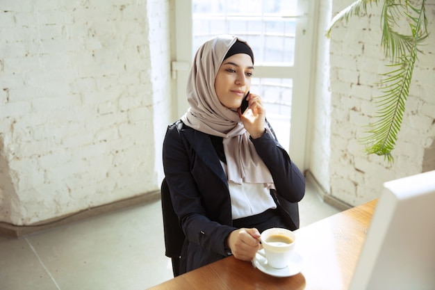 Muslim woman worker talking on phone while drinking coffee