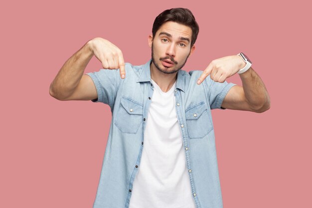 Must be here and right now. portrait of serious handsome bearded young man in blue casual style shirt standing, pointing down and looking at camera. indoor studio shot, isolated on pink background.