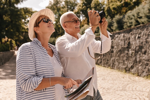 Free photo mustachioed man in glasses and light shirt photographing and smiling with blonde woman in hat and striped blouse outdoor.