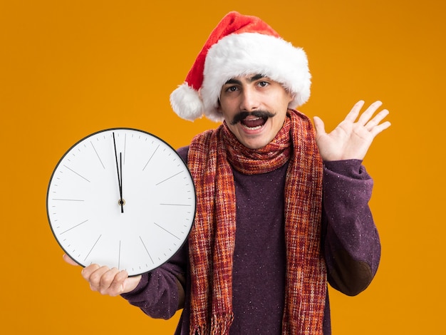 Mustachioed man wearing christmas santa hat with warm scarf around his neck holding clock  happy and excited  standing over orange wall