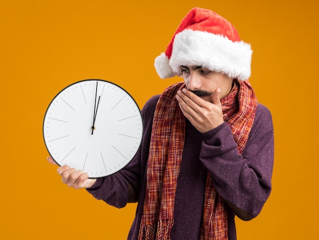 Uomo baffuto che indossa un cappello da babbo natale con una sciarpa calda intorno al collo tenendo l'orologio guardandolo stupito che copre la bocca con la mano in piedi sul muro arancione