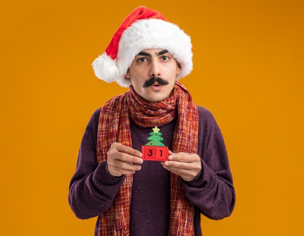 Усатый мужчина в рождественской шляпе санта-клауса с теплым шарфом на шее держит игрушечные кубики с новогодней датой, глядя в камеру, обеспокоенный стоя на оранжевом фоне