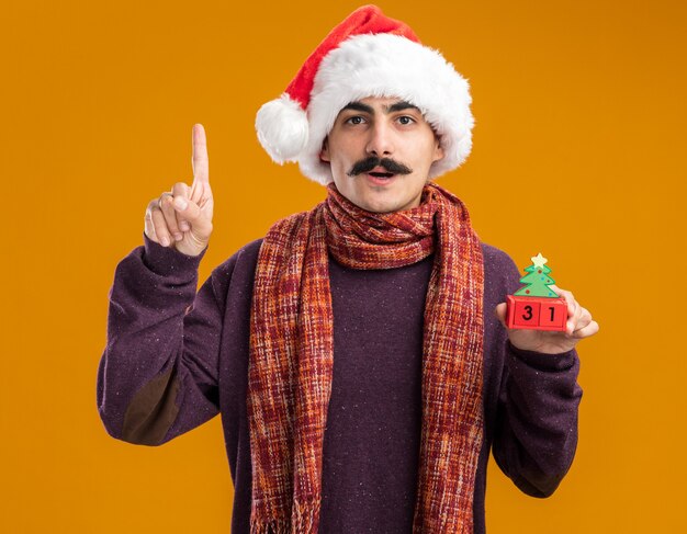 Усатый мужчина в рождественской шапке санта-клауса с теплым шарфом на шее держит игрушечные кубики с новогодней датой, выглядит уверенно, показывая указательный палец, стоящий на оранжевом фоне