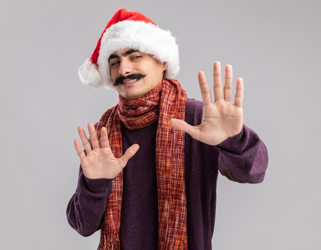 Mustachioed man wearing christmas santa hat with warm scarf around his neck looking confused making defense gesture with hands 
