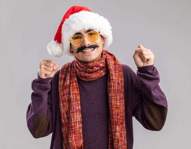 Mustachioed man wearing christmas santa hat and yellow glasses with warm scarf around his neck clenching fists happy and excited standing over white  wall