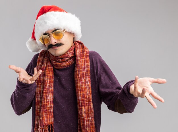 Baffuto, uomo, il portare, natale, cappello santa, e, occhiali gialli, con, caldo, sciarpa, intorno, suo, collo, guardando macchina fotografica, confuso, con, braccia fuori, standing, bianco, fondo