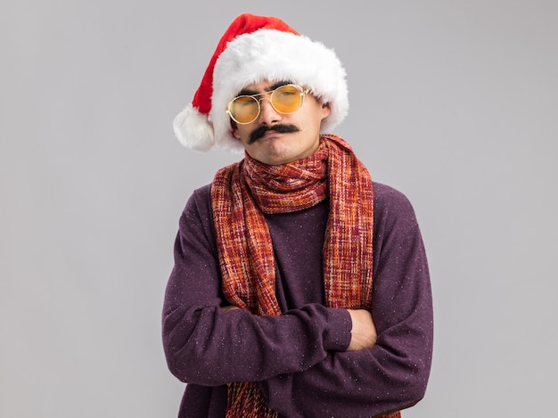 Free photo mustachioed man wearing christmas santa hat and yellow glasses with warm scarf around his neck  with serious face with arms crossed standing over white  wall