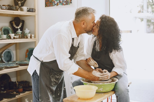 Mutual creative work. Adult elegant couple in casual clothes and aprons. People creating a bowl on a pottery wheel in a clay studio.