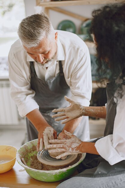 Mutual creative work. Adult elegant couple in casual clothes and aprons. People creating a bowl on a pottery wheel in a clay studio.