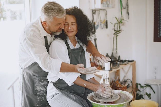 Mutual creative work. Adult elegant couple in casual clothes and aprons. People creating a bowl on a pottery wheel in a clay studio.