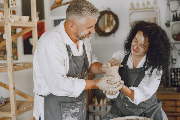 Mutual creative work. Adult elegant couple in casual clothes and aprons. People creating a bowl on a pottery wheel in a clay studio.