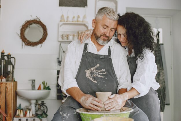 Mutual creative work. Adult elegant couple in casual clothes and aprons. People creating a bowl on a pottery wheel in a clay studio.