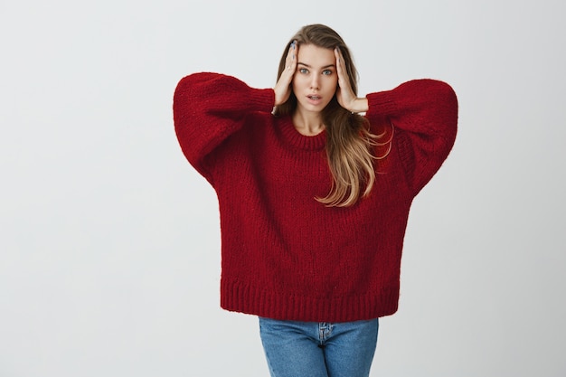 My head is spinning round. Portrait of expressive sensual woman with attractive appearance standing  in red loose sweater, holding hands on face and making model expression