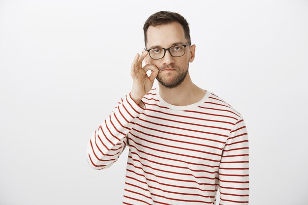 My mouth is fortress, count on me. Portrait of serious funny caucasian man in glasses, making gesture with hand, zipping lips