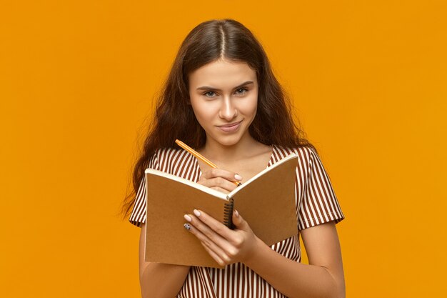 Mysterious attractive young female with long hair holding open copybook and pen writing down