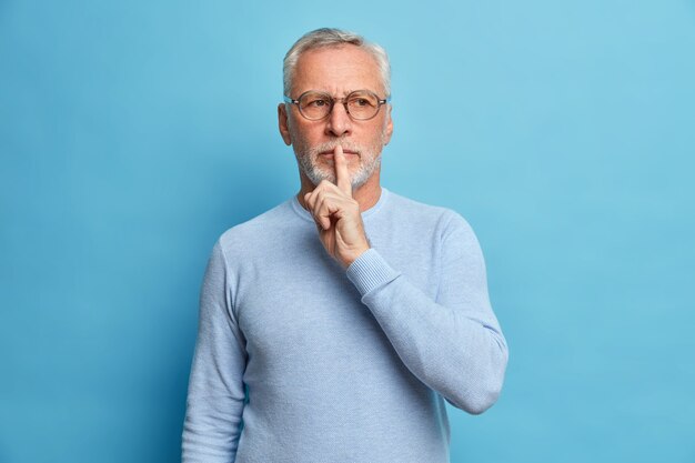 mysterious bearded man makes silence gesture has thoughtful expression asks to be quiet tells secret wears casual jumper and optical glasses isolated over blue wall