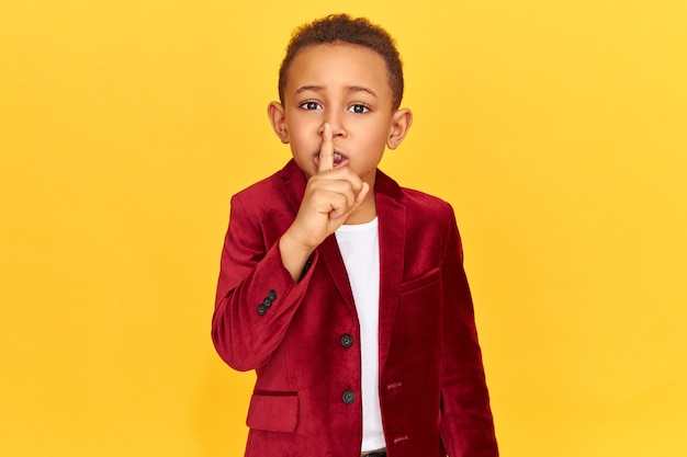 Mysterious boy in stylish clothes making shush gesture with fore finger at his lips