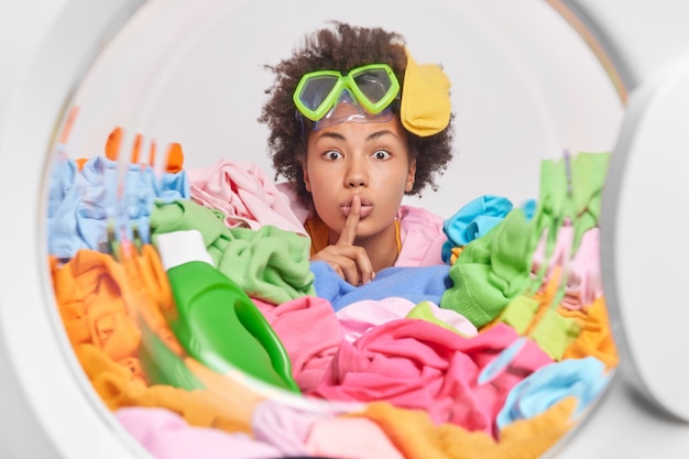 Free photo mysterious busy curly housekeeper makes hush gesture looks surprisingly at front stuck in dirty laundry wears snorkeling glasses poses in washing machine door