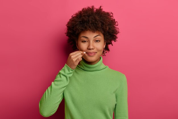 Mysterious curly woman zips mouth shut, tells secret, closes lips on lock, makes promise not tell anyone confidential information, wears green jumper