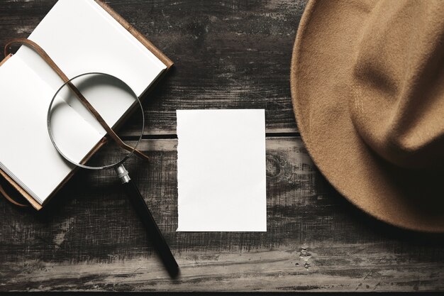 Mysterious detective game concept. Opened notebook in leather cover, sheet of white paper, felt brown hat and big vintage magnifier steel glasses isolated on black aged wood table. Top view.