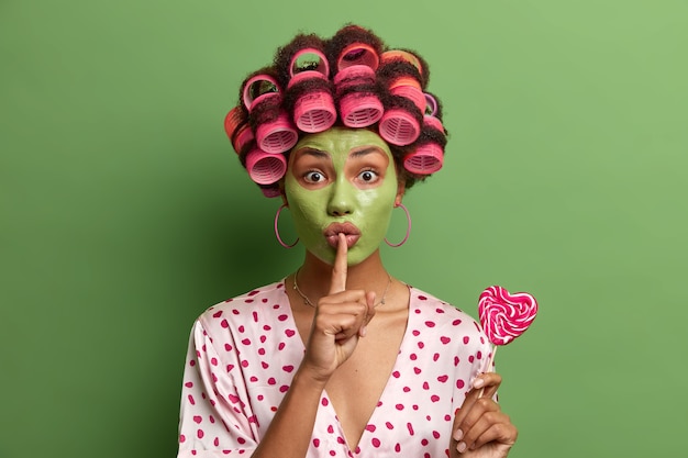 Mysterious ethnic woman wears green facial mask and hair rollers, makes silence gesture, tells secret, holds delicious heart shaped lollipop, makes hairstyle, wears dresssing gown, spends time at home