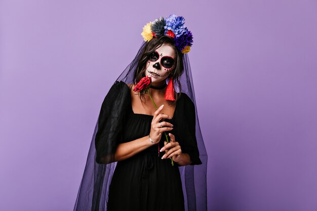 Mysterious Mexican woman in dress of black widow posing on lilac wall. Photo of girl with crown of flowers and bright earrings.