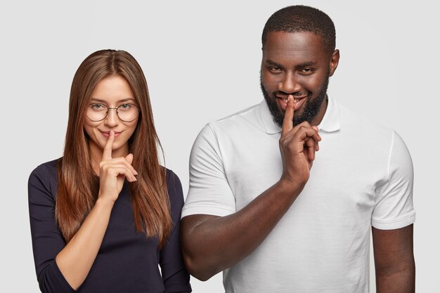 Mysterious multiethnic woman and man look secretly, keep both index fingers on lips as try be mute